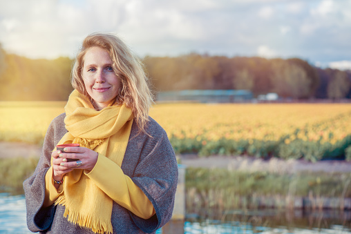 Portrait of a serene woman in  poncho and scarf holding a thermos cup with hot tea in the countryside. In the background  yellow tulip fields are situated.