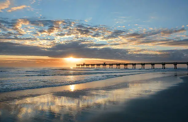 Sun rising over horizon and pier, beach illuminated with sunlight, beautiful sky reflected on the beach. Jacksonville Florida, USA.