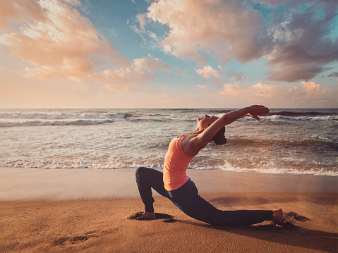 Vintage retro effect filtered hipster style image of Yoga outdoors - sporty fit woman practices yoga Anjaneyasana - low crescent lunge pose outdoors at beach on sunset