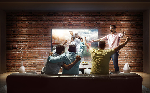 :biggrin:A group of young male friends are cheering while watching Soccer game at home. They are sitting on a sofa in the modern living room faced to a big TV set on the front wall.