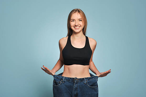 Fit young woman in loose jeans Fit young woman in loose jeans after losing weight isolated on blue background woman defeat stock pictures, royalty-free photos & images