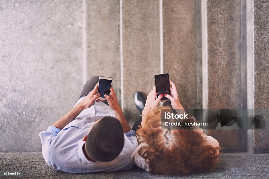 Young couple texting messages over smart phones Young man and woman are leaned on wall, standing on stairs, using their smart phones to text messages. Aerial view Two People Stock Photo