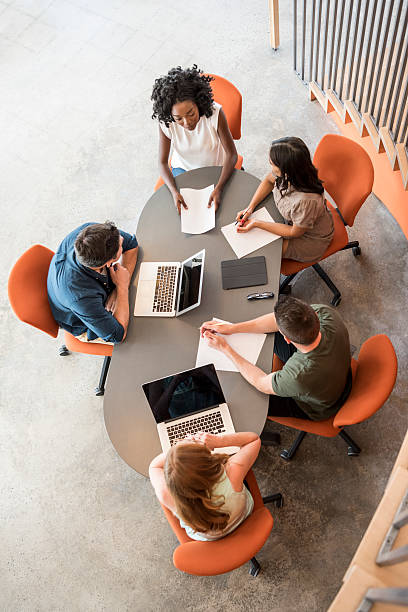Overhead view of five business people at table in meeting They are sitting on modern orange chairs with laptops and taking notes vertical stock pictures, royalty-free photos & images