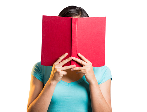 A young latin woman holding book over her face in a horizontal waist up shot with white background.