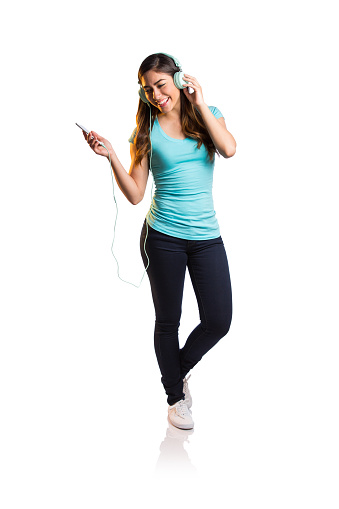 A young latin woman listening to music with headphones and smiling away in a vertical full length shot with white background.