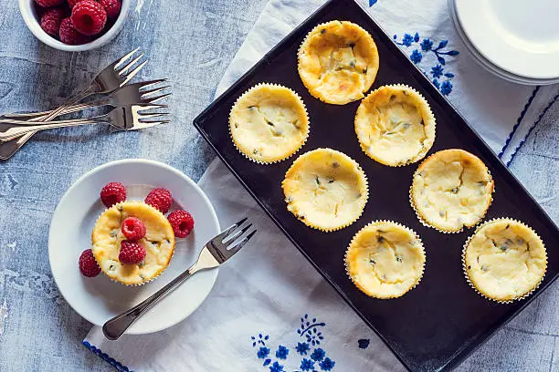 Mini passion fruit cheesecakes in cupcake liners served with fresh raspberries. The cheesecakes are in a metal tray resting on a floral napkin next to forks and plates.