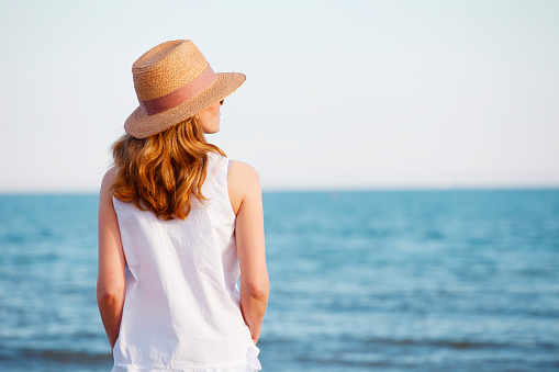 Rear view shot of a woman wearing casual clothes and straw hat while standing by the sea.