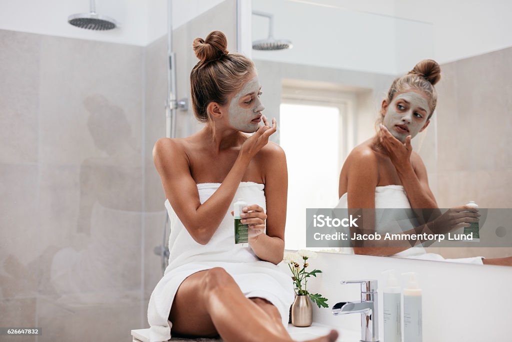 Young woman in bathroom applying facepack Young woman sitting in bathroom and applying facepack. Beautiful female in front of mirror applying facial mask. Mask - Disguise Stock Photo