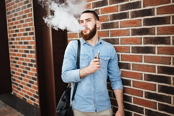 young man with beard vaping an electronic cigarette outdoor. - one person looking at camera male posing imagens e fotografias de stock