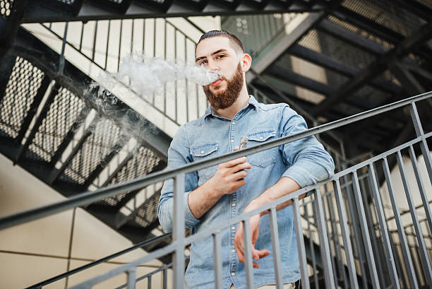 young man with beard vaping an electronic cigarette outdoor. - one person looking at camera male posing imagens e fotografias de stock