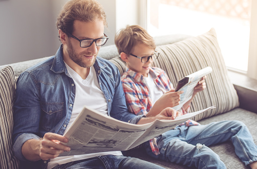 Father and son are reading newspapers and smiling while spending time together at home
