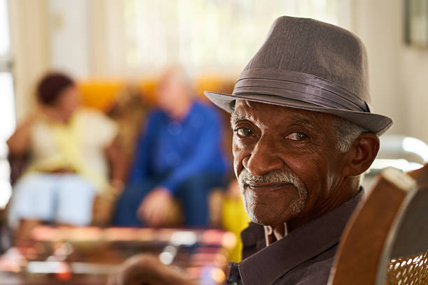 Senior Black Man With Hat Looking At Camera In Hospice Portrait of elderly black man looking at camera in retirement home, with group of friends in background. Patients relaxing in hospice for seniors. cuban ethnicity photos stock pictures, royalty-free photos & images