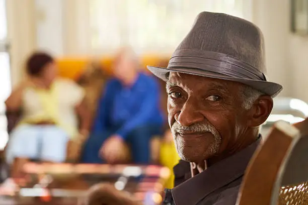 Portrait of elderly black man looking at camera in retirement home, with group of friends in background. Patients relaxing in hospice for seniors.