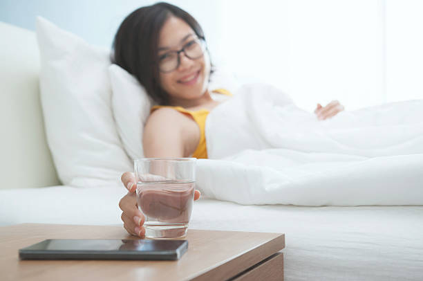 beautiful asian girl smiling and lying on bed. - transparent holding glass focus on foreground imagens e fotografias de stock