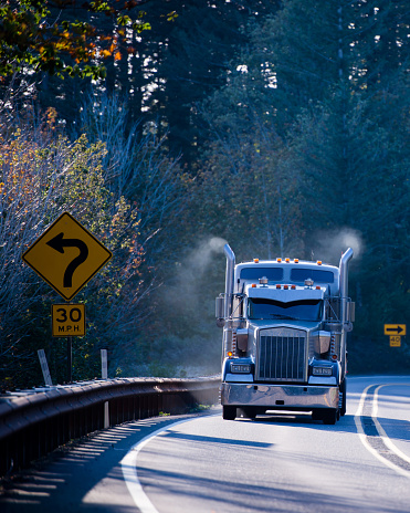 Huge blue classic big rig semi truck with chrome tailpipes highest of which is the smoke from the burned fuel the engine and semi trailer, turn on a winding road in the sunlight among the trees.