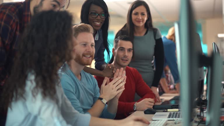 Closeup side view of group of software developers working late at IT office. There are 6 mixed race people gathered around a dual display computer. 4k video. Dolly shot.