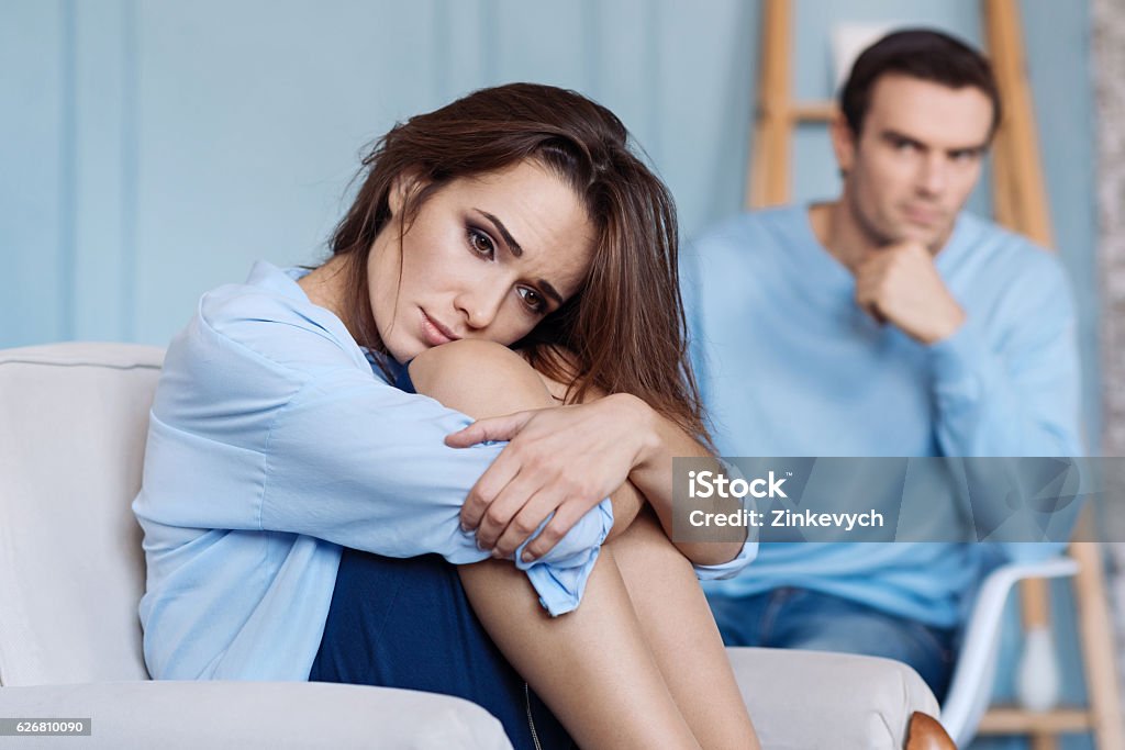 Moody yong woman sitting at home Forget all troubles. Gloomy thoughtful beautiful woman folding her legs and sitting in the arm chair while her husband sitting in the background Couple - Relationship Stock Photo