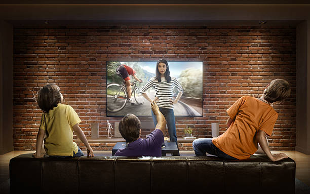 Family with children cheering and watching Cycle race on TV :biggrin:Father and two young children cheering and watching Cycle race on TV. Mother Stands in front of the TV. They are sitting on a sofa in the modern living room. The TV set is on the loft brick wall. It is evening outside the window. track and field fans stock pictures, royalty-free photos & images