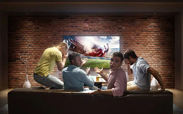 :biggrin:A group of young male friends are cheering while watching Baseball game at home. They are sitting on a sofa in the modern living room faced to a big TV set on the front wall.