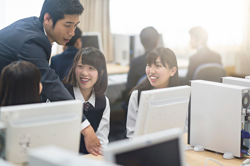 Teacher assisting high school girls in computer class. Smiling female students are sitting in brightly lit school. They are wearing uniforms.