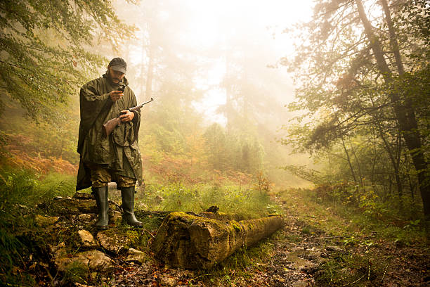 Hunter in the forest Portrait of hunter with rifle and satellite phone in the forest. There is a fallen tree next to him. woodland camo stock pictures, royalty-free photos & images