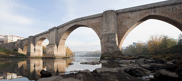 Panoramic image of the Roman Bridge (Ponte Vella) in Ourense taken in morning light in late autumn. Other bridges are visible through the main archway. River rocks appear in the foregroundand the calm waters of the Miño are present.