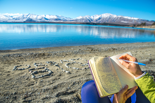 Happy Young woman surrounded by mountains and nature writing a diary on the shore of Lake Tekapo, New-Zealand