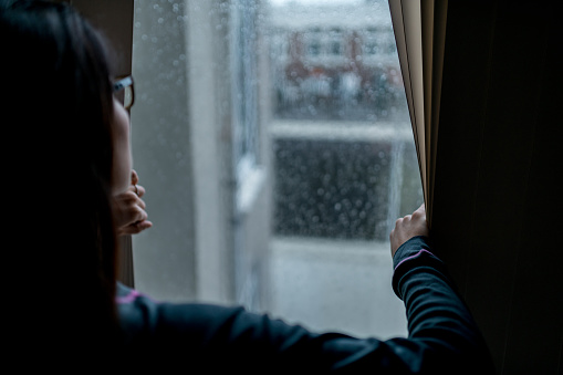 Woman standing next to the window ,holding a curtain and looking outside