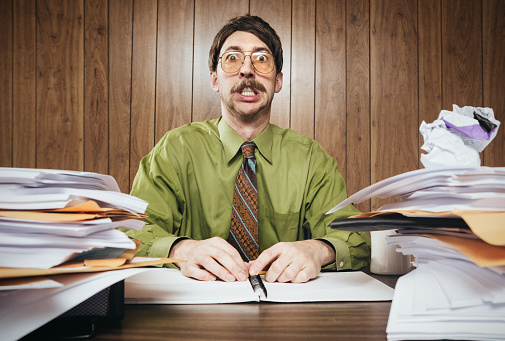 A white collar business man working in a retro 1980's style office sits at a desk piled with paperwork and documents.  He looks tired and overwhelmed, or surprised at all the work he has to do.  Horizontal image.