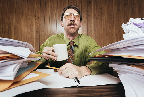 A white collar business man working in a retro 1980's style office sits at a desk piled with paperwork and documents.  He looks tired and overwhelmed at all the work he has to do.  Horizontal image.