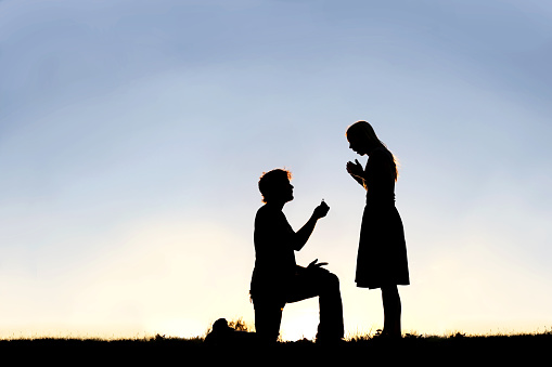 A silhouette of a young man, down on one knee and holding a diamond engagement ring, proposing to his girlfriend.