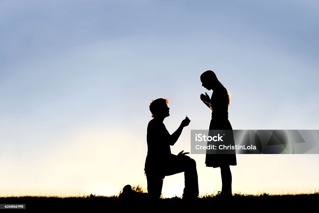 Silhouette of Young Man with Engagement Ring Proposing to Woman A silhouette of a young man, down on one knee and holding a diamond engagement ring, proposing to his girlfriend. Engagement Stock Photo