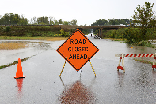 A bright orange Road Closed Ahead Sign and caution cones and barracades are blocking a rain flooded rural street.