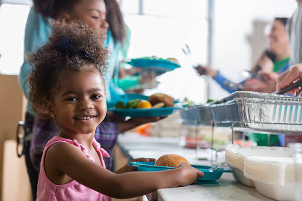 Adorable little girl in soup kitchen Cheerful African American little girls smiles while in line in a soup kitchen. She is holding a plate full of healthy food. Her family is in line behind her. soup kitchen stock pictures, royalty-free photos & images