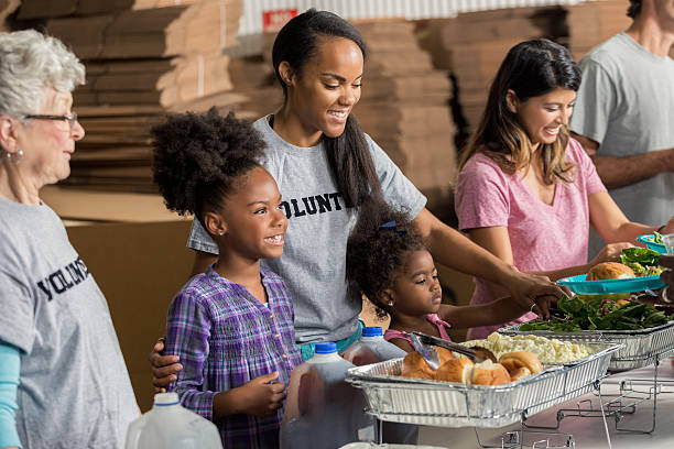 Diverse family volunteers together in soup kitchen African American mother and her two daughters serve meals to the homeless with their diverse family. soup kitchen stock pictures, royalty-free photos & images