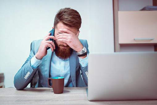Photo of a young frustrated businessman with his hands on his face, he is exhausted and worried about his work problems. Talking on his mobile phone in front og his laptop computer and coffee mug.