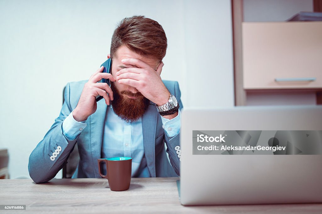 Worried and Exhausted Businessman Talking on Phone in his Office Photo of a young frustrated businessman with his hands on his face, he is exhausted and worried about his work problems. Talking on his mobile phone in front og his laptop computer and coffee mug. Using Phone Stock Photo