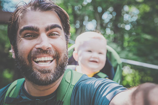 DSLR Picture of a cheerful father Backpacking or Hiking with a Baby boy inside a baby carrier in Forest on a nice day of summer. The father is taking a selfie and smiling.