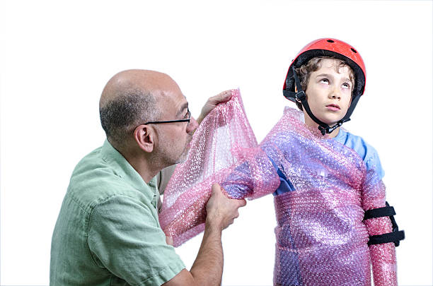 Overprotecting dad putting plastic bubbles over kid before skatebording Overprotecting dad putting plastic bubbles over kid before skatebording elbow pad stock pictures, royalty-free photos & images