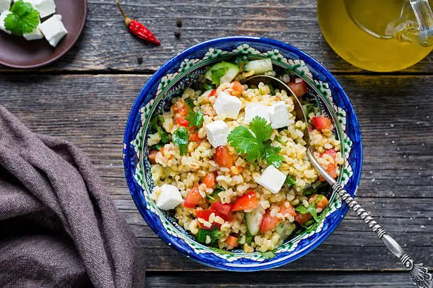 Bowl of healthy and delicious fresh tabbouleh salad with bulgur and feta cheese on wooden table background, top view