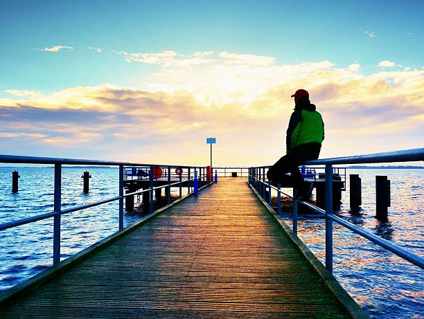 Man in warm jacket and baseball cap sit on pier and enjoy quiet morning sea. Tourist relax. Sunny clear blue sky, smooth water level