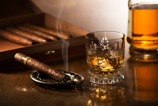 Glass of whiskey with ice cubes and smoking cigar on wooden table