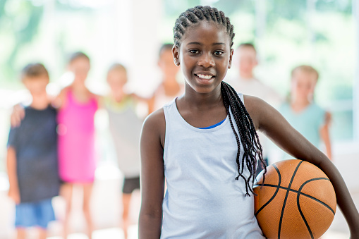 A multi-ethnic group of elementary age children are playing together at the gym during recess. One girl is holding a basketball and is smiling while looking at the camera.