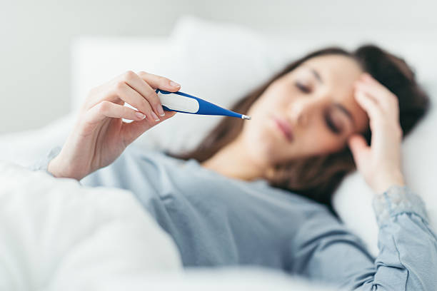 Woman measuring her temperature Woman with flu virus lying in bed, she is measuring her temperature with a thermometer and touching her forehead fever stock pictures, royalty-free photos & images