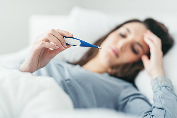 Woman measuring her temperature Woman with flu virus lying in bed, she is measuring her temperature with a thermometer and touching her forehead temperature stock pictures, royalty-free photos & images