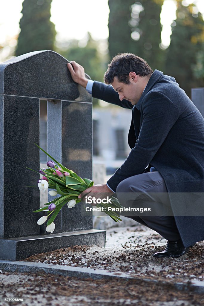 They were her favorite flowers Shot of a young man visiting a gravesite with a bunch of flowers Funeral Stock Photo