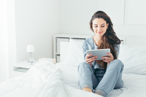 Young smiling woman sitting in the bed at home and connecting with a tablet, relax and leisure concept