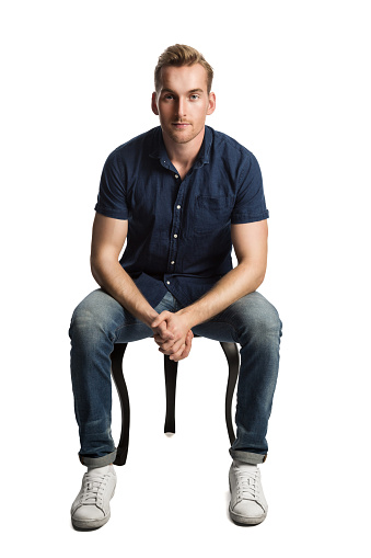A sad man sitting down on a stool in front of a white background, wearing a blue shirt and jeans with white shoes, looking at camera.