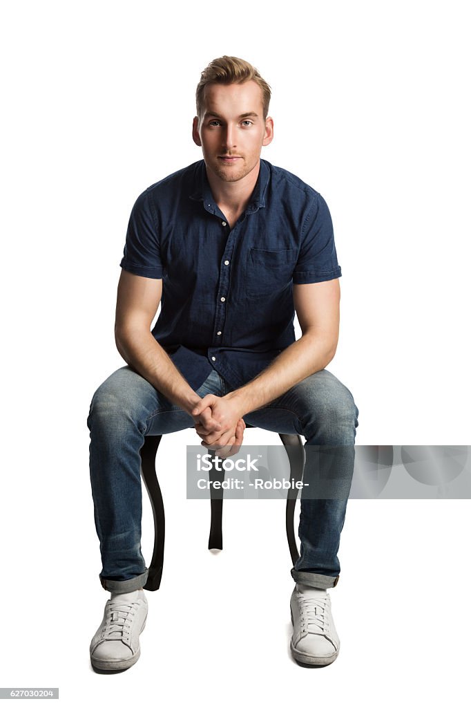 Man in blue shirt and jeans sitting down A sad man sitting down on a stool in front of a white background, wearing a blue shirt and jeans with white shoes, looking at camera. Sitting Stock Photo