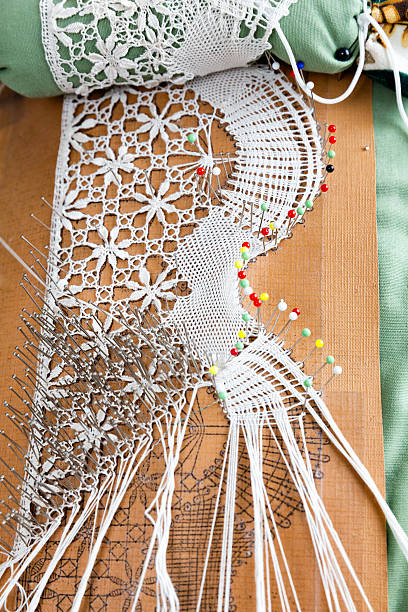 Bobbin lace labour Bobbin lace labour  is also known as pillow lace, because it was worked on a pillow, and bone lace, because early bobbins were made of bone.It is a lace textile made by braiding and twisting lengths of thread, which are wound on bobbins to manage them. As the work progresses, the weaving is held in place with pins set in a lace pillow, the placement of the pins usually determined by a pattern or pricking pinned on the pillow. lacemaking photos stock pictures, royalty-free photos & images